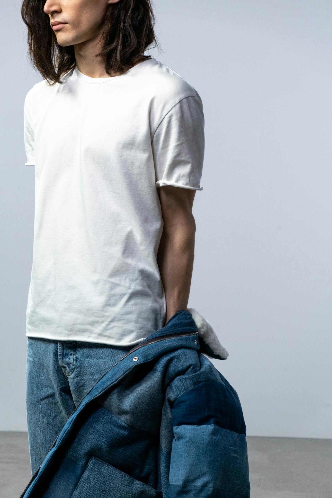 A man wearing a white shirt and denim jeans holds a jacket, posing in a studio setting.
