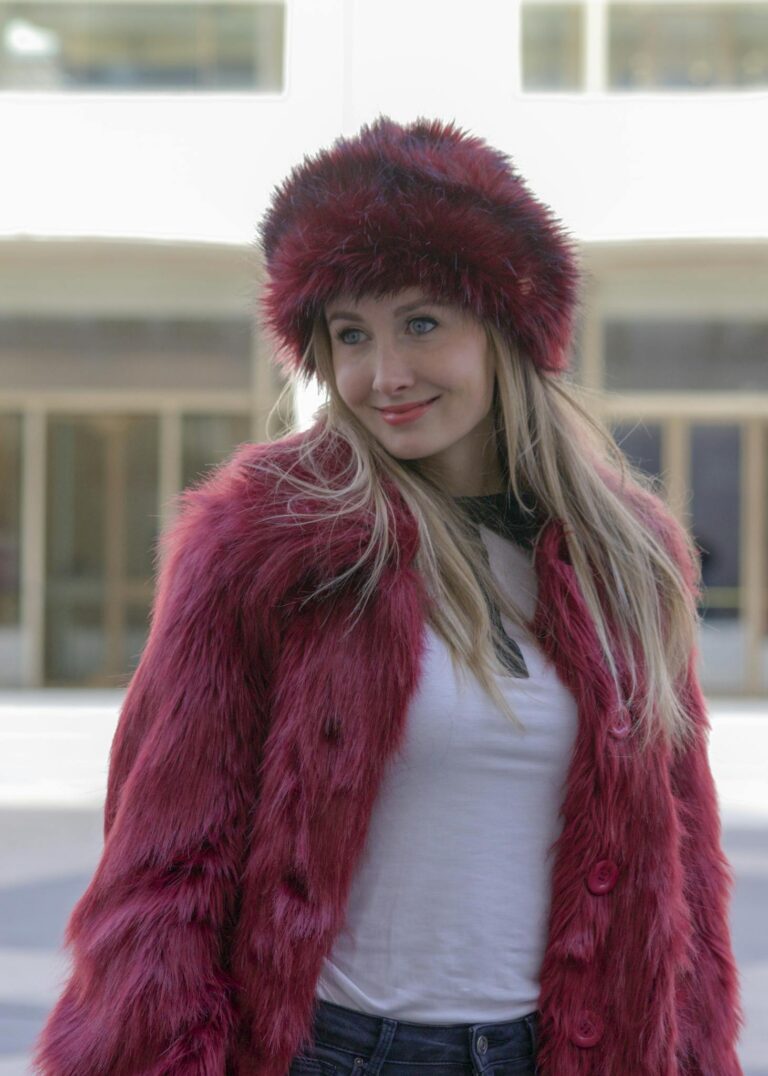 Stylish woman wearing a red fur hat and coat, casually standing in a city environment.