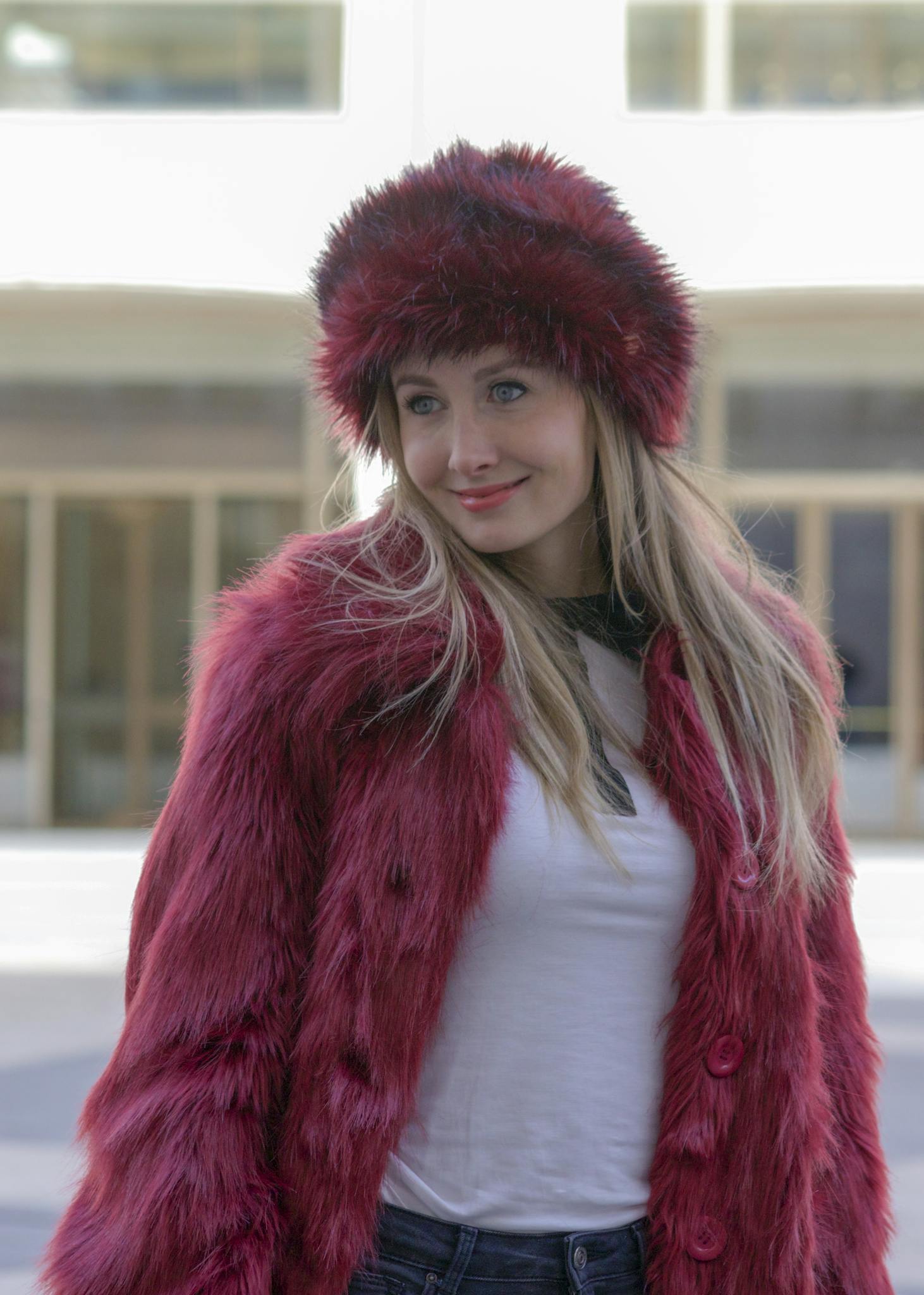 Stylish woman wearing a red fur hat and coat, casually standing in a city environment.