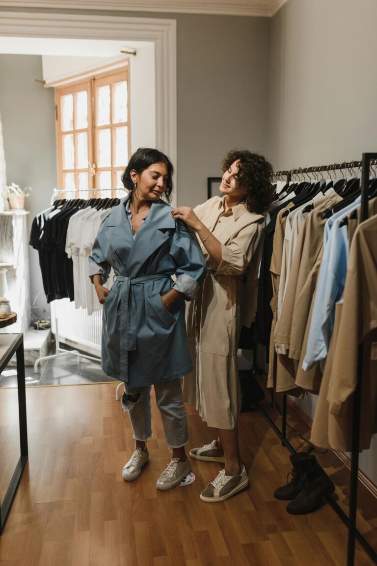 Two women shopping in a boutique, trying on stylish trench coat outfits.